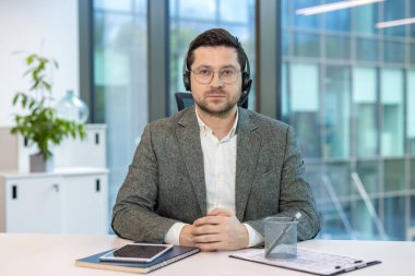 Kulaklık ve takım elbiseli gülümseyen genç bir adamın yakın plan portresi ofiste oturmuş kameraya el sallıyor ve konuşuyor..