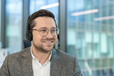 Kulaklık ve gözlük takan genç bir adamın yakın plan fotoğrafı. Ofiste çalışıyor, gülümsüyor ve yan tarafa bakıyor..