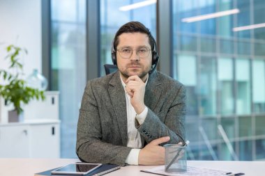 Takım elbiseli ve kulaklıklı ciddi bir gencin portresi masasında oturmuş, kendine güvenerek kameraya bakıyor..