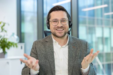 Kulaklık takmış gülümseyen genç bir adamın portresi. Ofis masasında oturuyor ve elleriyle kameraya konuşuyor..