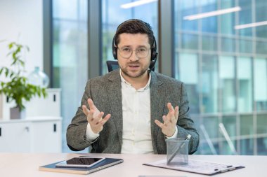 Kulaklık ve bilgisayar takmış genç bir adamın portresi bir masada oturuyor ve bir online video konferansı düzenliyor, elleriyle işaret ederek anlatıyor..
