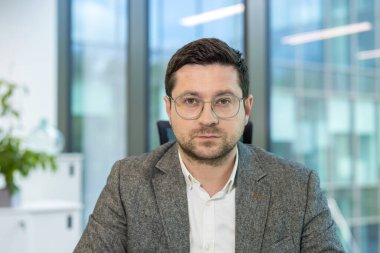 Ofisinde oturmuş, kameraya ciddi ciddi bakan genç bir erkek işadamının portresi..