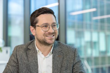 Gözlüklü, gülümseyen bir adam, parlak, modern bir ofis binasının önünde oturuyor..