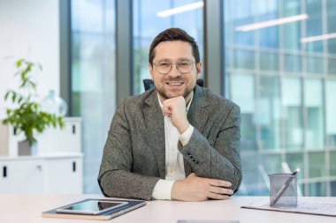 Ofiste takım elbiseli, gözlüklü, gülümseyen ve kameraya bakan genç bir işadamının portresi..