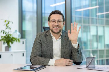 Ofisindeki işyerinde oturmuş gülümseyen genç adam, kameraya el sallıyor ve selamlıyor..