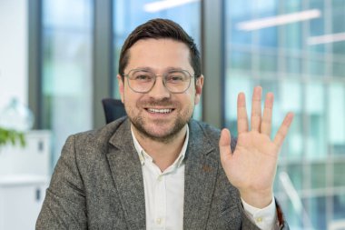 Kameraya el sallayan gülümseyen bir işadamı profesyonel bir ortamda çekilmiş dostane bir jest..