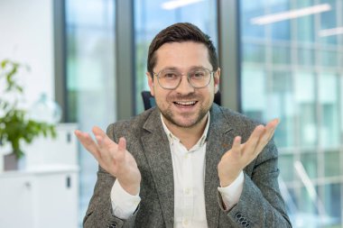 Takım elbiseli ve gözlüklü genç bir işadamı ofiste oturuyor ve elleriyle kameraya konuşuyor..