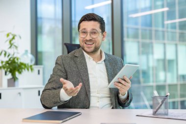 Bir iş adamı video konferansında elinde tablet ve ofis ortamında el kol hareketi yapıyor. Sanal bir toplantıda gibi görünüyor.