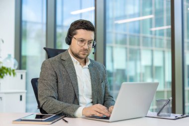 Takım elbiseli ve kulaklıklı ciddi bir genç ofiste bir masada otururken dizüstü bilgisayarla çalışıyor..