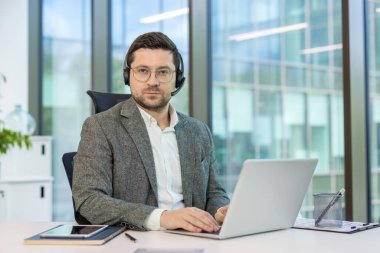 Ciddi bir genç adam, ofis çalışanı ve danışman masada oturuyor, dizüstü bilgisayarda çalışıyor ve kameraya bakıyor..