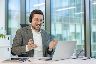 Gözlüklü ve kulaklıklı bir işadamı, bilgisayar ekranına bakarken el hareketleriyle bir video konferansına katılıyor..