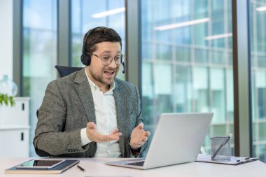 Takım elbiseli ve kulaklıklı genç bir adam dizüstü bilgisayarında bir video görüşmesi yapıyor, satıyor, açıklıyor ve elleriyle işaret ediyor..