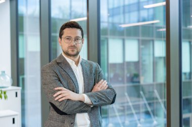 Ofiste cam kenarında duran takım elbiseli ve gözlüklü ciddi bir genç işadamının portresi, kolları göğsünde ve kameraya güvenle bakıyor..