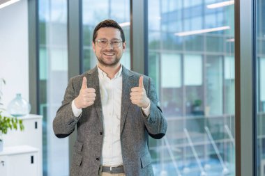 Gülümseyen genç adam, takım elbiseli ve gözlüklü bir ofis çalışanı ofiste durmuş kameraya bakıyor ve parmaklarıyla süper bir işaret gösteriyor..