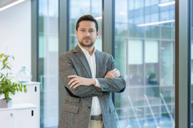 Ofiste takım elbiseli ve kravatlı genç ve başarılı bir işadamı kolları göğsünde sıkıca kameraya bakıyor..