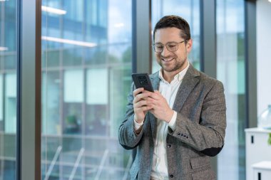 Gülümseyen bir işadamı, gözlük takıyor, akıllı telefon kullanıyor, modern ofis ortamında büyük bir pencerenin yanında duruyor..