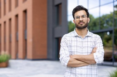 Ciddi bir ifadeyle Hintli bir adam kollarını kavuşturmuş modern bir binanın önünde duruyor..