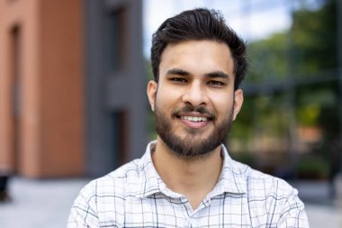 Genç, gülümseyen bir öğrenci kameraya poz veriyor, kendinden emin ve arkadaş canlısı bir ifade sergiliyor, açık havada bir binayla..