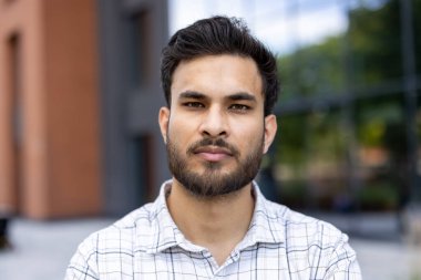 Odaklanmış bir Hintli doğrudan kameraya bakar, yakın çekimde görüntülenir. Dışarıda kareli bir gömlek giyiyor..