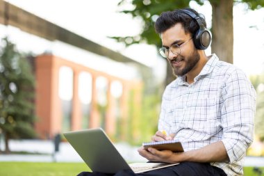 Dışarıda dizüstü bilgisayarda çalışan, kulaklık takan ve deftere yazı yazan genç bir adam. Arka planda ağaçlar ve bir bina görünüyor..