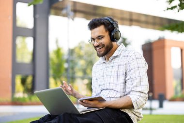 Dışarıda bankta oturan, kulaklık takan, elinde kalemle not defteri tutan ve video konuşmasında gülümseyen genç Hintli adam..