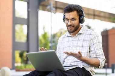 Genç bir öğrenci kulaklık takıyor ve dışarıdaki bir video çağrısına katılıyor, bilgisayarını arkadaşça bir ifadeyle kullanıyor..