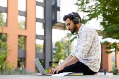 Kulaklık takan genç bir adam laptopuyla dışarıda, modern bir binanın yanında çalışıyor..