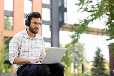 Kulaklık takan genç bir öğrenci güneşli bir günde laptopuyla dışarıda çalışıyor..