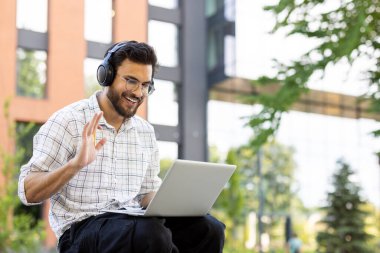 Dışarıda dizüstü bilgisayar kullanırken kulaklık takan, gülümseyen bir adam bir video çağrısında görünüyor..