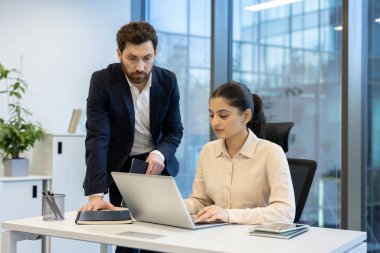 İki meslektaş modern ofis ortamında işbirliği yapıyor, biri dizüstü bilgisayarda çalışırken diğeri dikkatle izliyor..