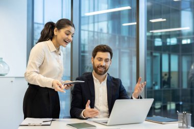 İki iş arkadaşı modern ofis ortamında iş birliği yapıyor, bir dizüstü bilgisayardaki verileri analiz ediyor, planları tartışıyor ve bir başarıyı kutluyorlar..