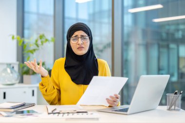 Gözlüklü Müslüman bir kadın iş aletleriyle çevrili bir ofiste belgelere bakar..