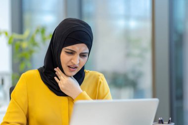 Başörtüsü takan bir kadın muhtemelen ofis ortamında dizüstü bilgisayarla çalışırken boğaz ağrısı çeker..