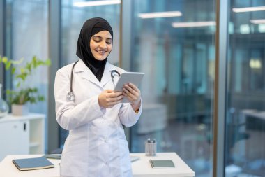Tesettürlü gülümseyen bir kadın doktor modern ofis ortamında bir tableti inceliyor..