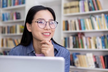 Gözlüklü bir Asyalı kadın gülümsüyor, çenesini elinde dinlendiriyor, kütüphane ortamında..