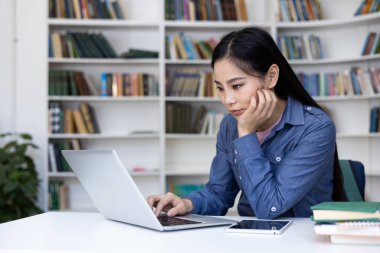Odaklanmış Asyalı bir kadın, kitaplarla ve dijital tabletlerle çevrili bir kütüphanede dizüstü bilgisayarla çalışıyor..