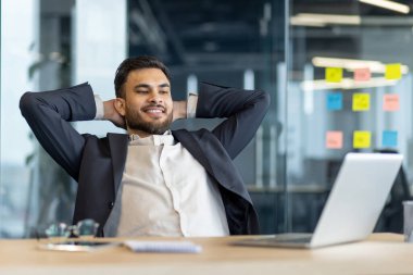 Takım elbiseli, gülümseyen bir işadamı ofisinde rahatlar, arkasını yaslanır, işinden zevk alır..