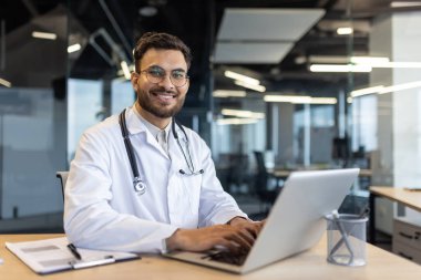 Beyaz önlük ve steteskop giyen gülümseyen bir doktor modern ofis ortamındaki bir masada dizüstü bilgisayarla çalışıyor..