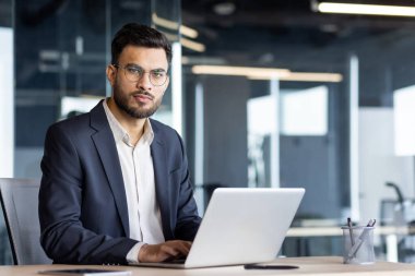 Donanma takım elbiseli ve gözlüklü, modern ofis ortamında dizüstü bilgisayarla çalışan kendine güvenen bir iş adamı..