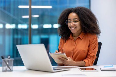 Gülümseyen genç bir kadın ofis ortamındaki bir video görüşmesi sırasında not alır. Laptopunu kullanıyor ve gözlük takıyor..