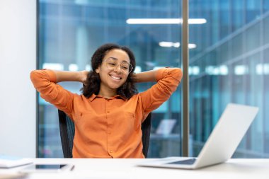 Afro-Amerikalı bir kadın kollarını başının arkasında dinlendirirken gülümsüyor. Ofis ortamında gözlük ve turuncu gömlek takıyor..