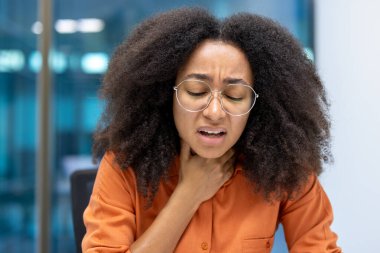 Endişeli bir kadın boğazını sıkıyor, muhtemelen rahatsızlığı ya da ağrısı var, ofis geçmişi olan bir evde..