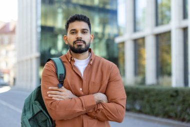 Genç, sakallı bir adam kolları bağlı, düğmeli bir gömlek ve sırt çantası takıyor. Arka planda modern bir bina var..