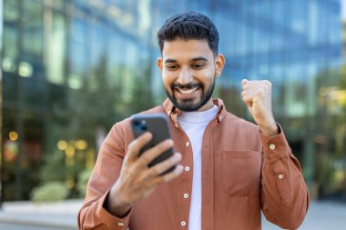 Genç Hintli bir adam telefonuna bakarken heyecanla gülümsüyor, modern bir binanın önünde başarıyı kutluyor..