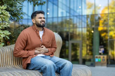 Açık havada oturan bir adam, midesini tutar, rahatsızlık gösterir, potansiyel bir hastalık ya da acı olduğunu gösterir. Sahnede bir bank ve arka planda modern bir bina yer alıyor..