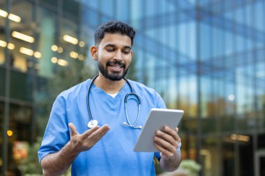 Önlüklü gülümseyen bir erkek doktor modern bir binanın önünde tablet kullanıyor, muhtemelen bir video görüşmesinde. Elleriyle hareket ediyor.
