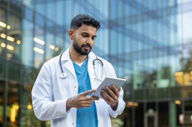 Laboratuvar önlüğü giyen bir doktor modern bir tıp binasının dışındaki tableti inceliyor. Boynunda steteskop var..