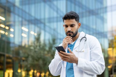 Endişeli bir doktor dışarıda telefonunu inceliyor, muhtemelen biriyle iletişim kuruyor ya da araştırıyor..