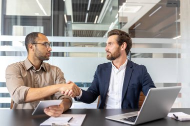 İki işadamı modern ofis ortamında el sıkışıyor, muhtemelen başarılı bir anlaşma ya da anlaşmadan sonra. İşbirliği ve takım çalışmasına odaklan.