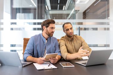 Modern bir ofiste iki farklı işadamı proje detaylarını dizüstü bilgisayarlar ve belgelerle tartışıyor..